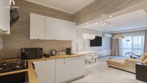 Cozinha moderna com fogão a gás, micro-ondas e armários brancos. Há uma área de jantar com mesa e cadeiras, e um sofá na sala de estar.
