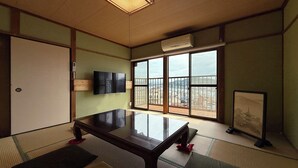 Quarto japonês tradicional com tatames, uma mesa baixa, uma TV e grandes janelas com vista para a cidade.