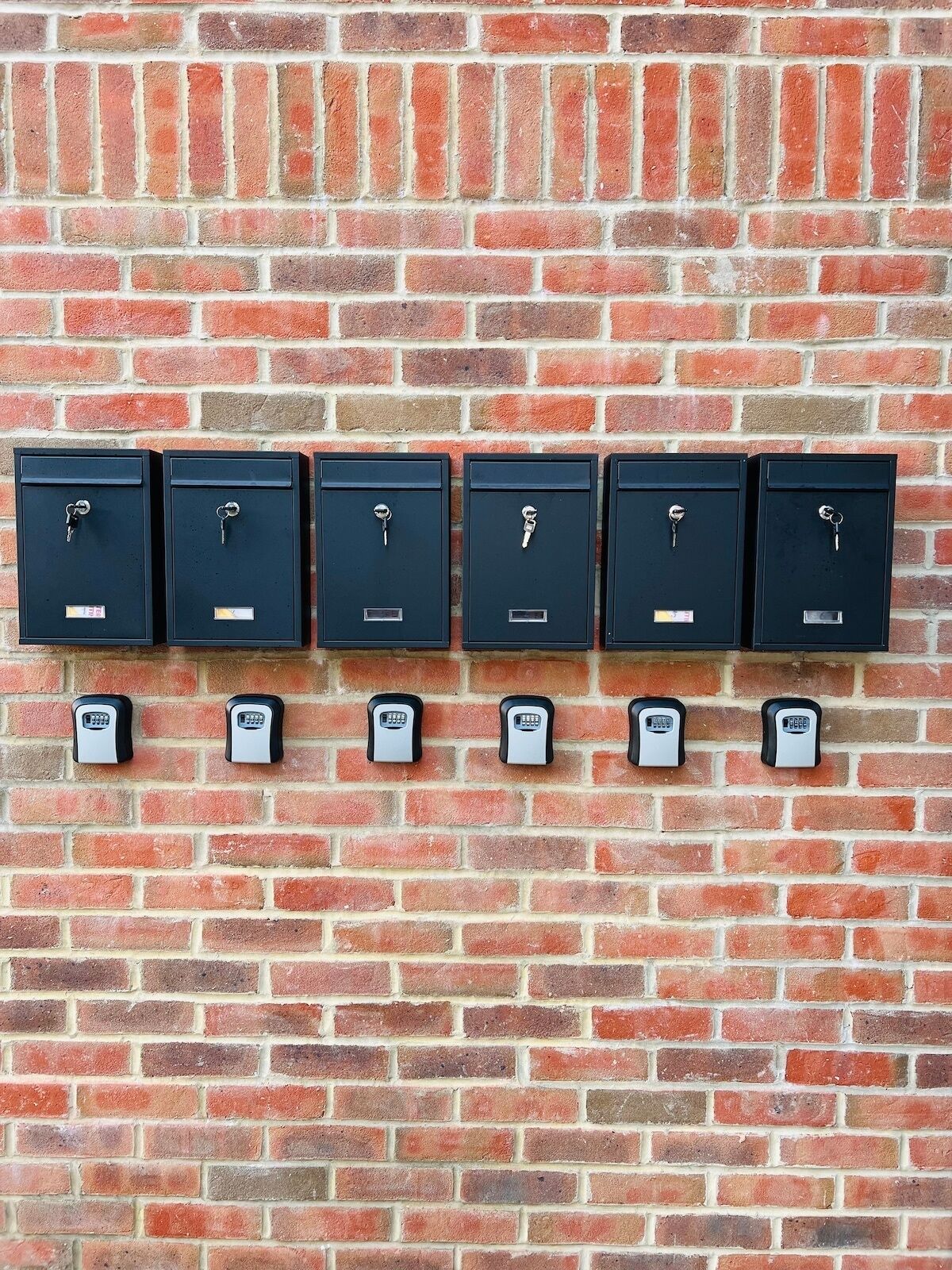 A row of six black metal mailboxes with keys and a keypad lock, mounted on a red brick wall.