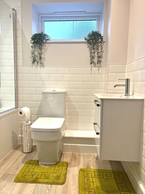 Une salle de bain moderne avec des toilettes, un lavabo et une baignoire. On y trouve deux tapis verts sur le sol, une fenêtre au-dessus de la baignoire et une plante sur le rebord de la fenêtre.