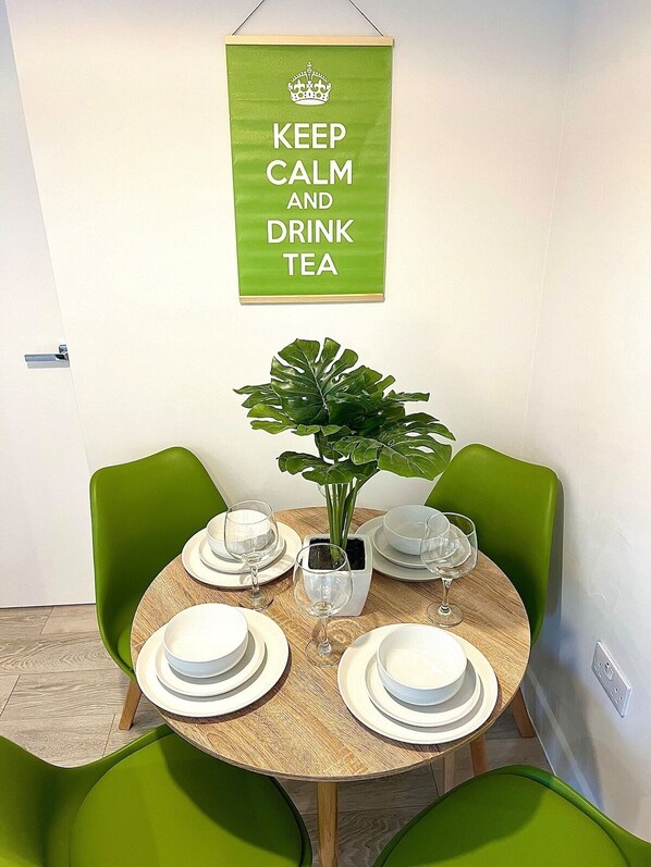 Un coin repas avec une table ronde en bois, dressée pour quatre personnes, entourée de chaises vertes. Un pot de fleurs est placé au centre, et une pancarte au-dessus indique : « GARDONS NOTRE CALME ET BUVONS DU THÉ ».