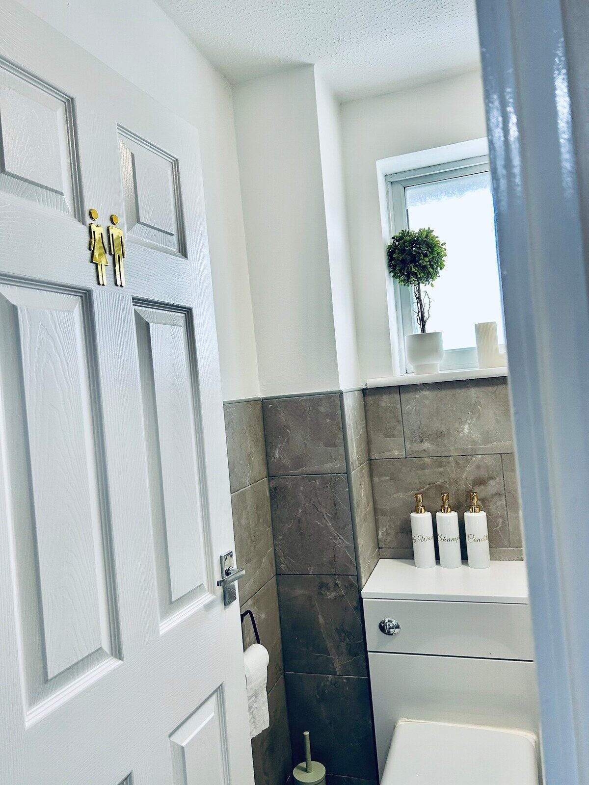 A bathroom with a white door, dark tiled walls, a small window with a plant, and three white bottles labeled "Shampoo" and "Conditioner" on a shelf.