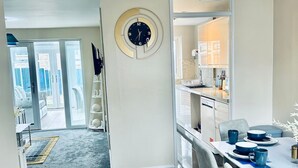 A modern kitchen with a round clock, a dining area with a blue pendant light, and a view through an open door to a patio. - Holiday Home with Parking , Near Colchester Zoo (Essex)