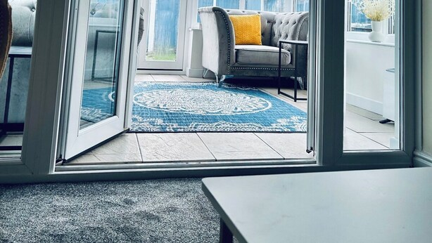 A conservatory with a glass door, a grey sofa with a yellow cushion, and a patterned rug. - Holiday Home with Parking , Near Colchester Zoo (Essex)