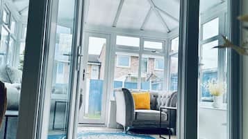 A conservatory with a glass door, a grey sofa with a yellow cushion, and a patterned rug.
