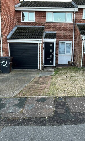 Une maison à deux étages, construite en briques, avec un garage, une porte d’entrée et un petit jardin à l’avant.