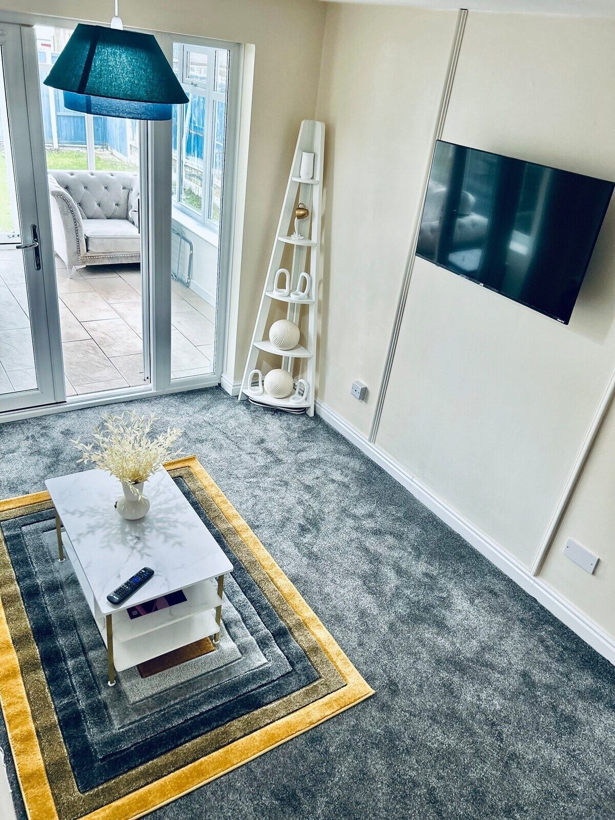 Una sala de estar con alfombra gris, una mesa de centro blanca con un florero, un televisor montado en la pared y un zapatero blanco.
