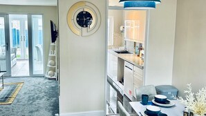 A modern kitchen with a dining area, featuring a clock, pendant light, and a view into the adjacent living space. - Holiday Home with Parking , Near Colchester Zoo (Essex)