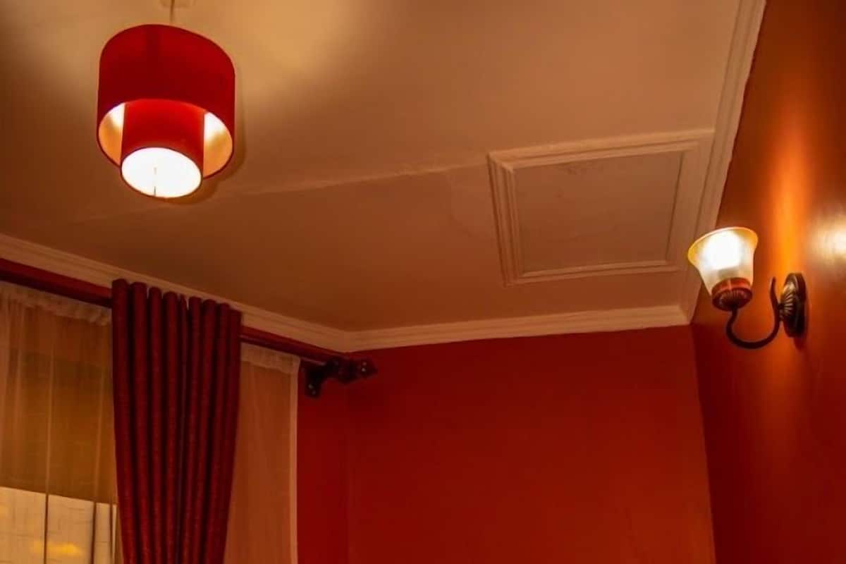 A hotel room with a bed, a window with curtains, a wall-mounted light fixture, and a ceiling light.