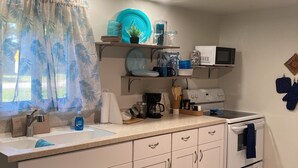 A kitchen with white cabinets, a sink, a stove, and a microwave. There are shelves above the counter holding dishes and a plant. - Lovely 2-bedroom mobile home with AC in Okeechobee, on the canal near the locks. (Okeechobee)