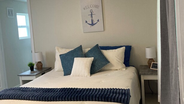 A neatly made bed with a white and blue color scheme, a welcome sign with an anchor, and a modern light fixture. - Lovely 2-bedroom mobile home with AC in Okeechobee, on the canal near the locks. (Okeechobee)
