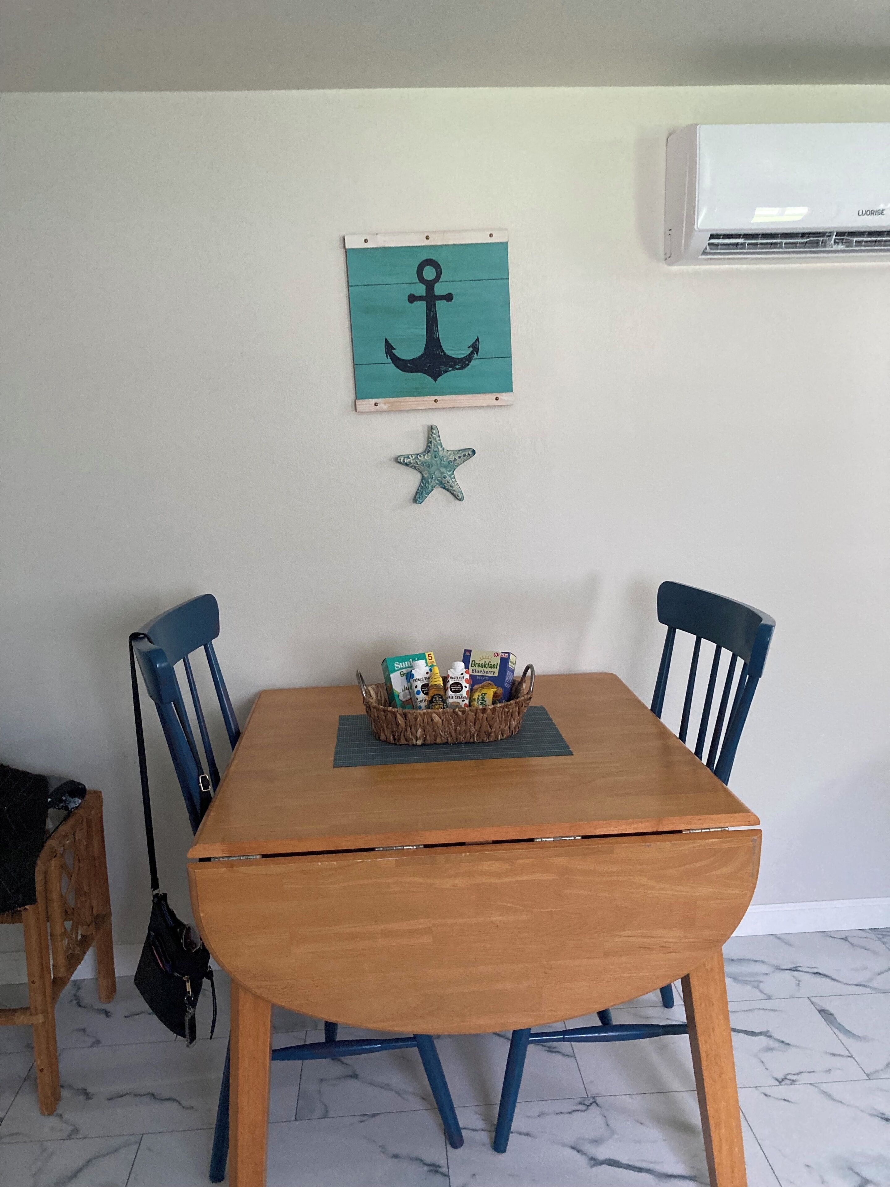 Una mesa de madera con una cesta tejida que contiene alimentos para el desayuno, flanqueada por dos sillas azules.