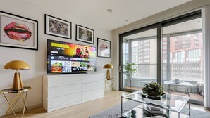 Una sala de estar moderna con un televisor de pantalla plana mostrando una película, un sofá, una mesa de centro y un gran ventanal con vistas a edificios.