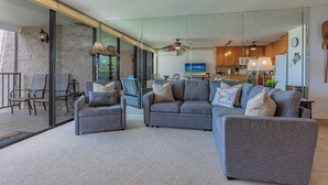 A modern living room with a sofa set, a dining area with chairs, a kitchen with wooden cabinets, and a large window.