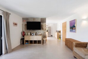 Un salon moderne avec un mur d’accent en pierre, un téléviseur à écran plat, une console en bois, un canapé en osier et une cuisine en arrière-plan.