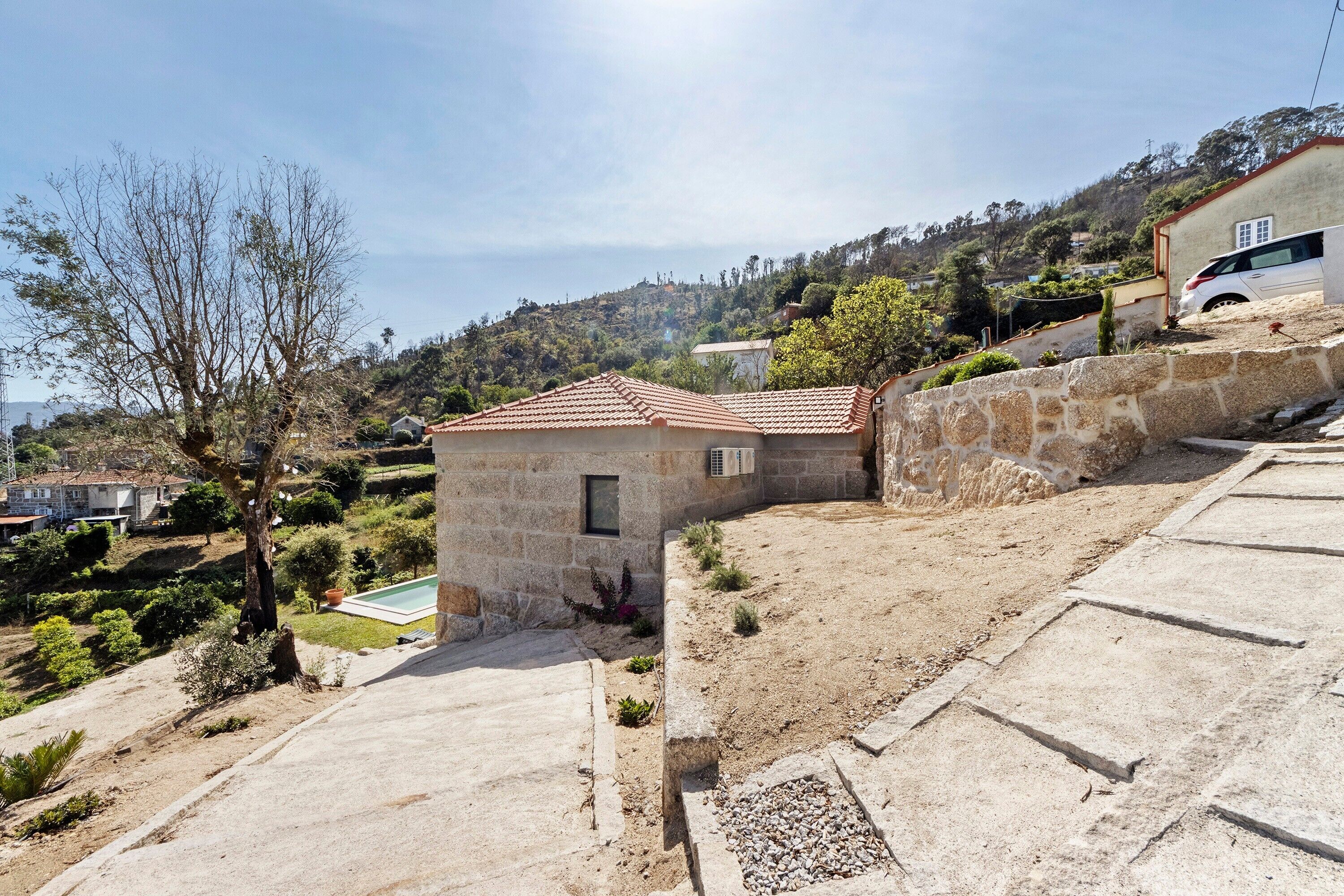 Una propiedad residencial con un muro de piedra, un edificio pequeño con techo de tejas y una zona pavimentada con piscina.