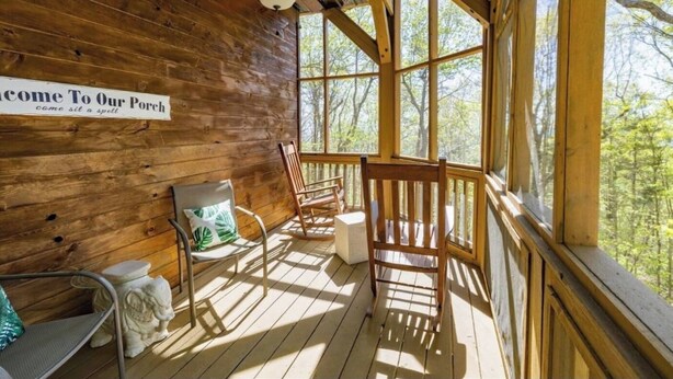 A wooden porch with a sign reading "Welcome to Our Porch," wooden chairs, and a table with a cushion.