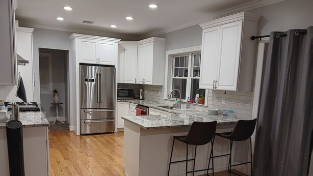 A modern kitchen with white cabinetry, a stainless steel refrigerator, a microwave, and a sink. There are two bar stools at the island. - Walking distance Golf Course 4-bedroom house with Living  Dining and Family Room (Farmingdale)