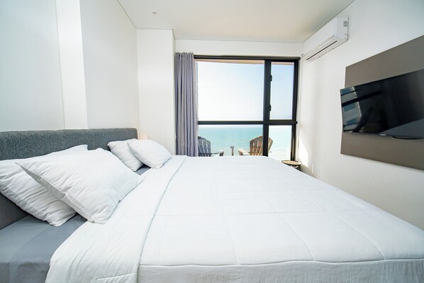 Une chambre d’hôtel avec un grand lit, un téléviseur fixé au mur et une vue sur la mer par la fenêtre.