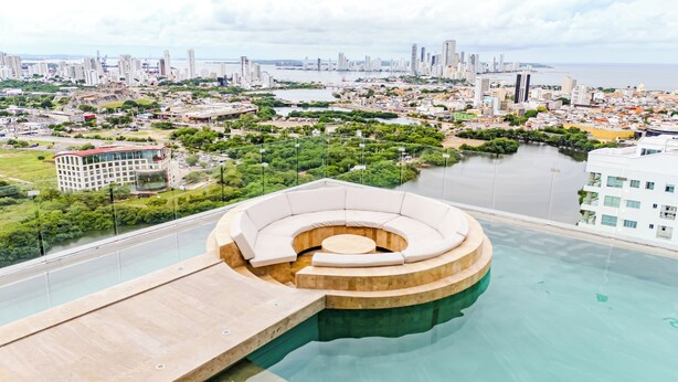 A rooftop infinity pool with a circular seating area, overlooking a cityscape with a river and high-rise buildings.