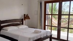 A hotel room with a bed, a bedside lamp, an air conditioner, and a sliding glass door leading to a balcony with a hammock.