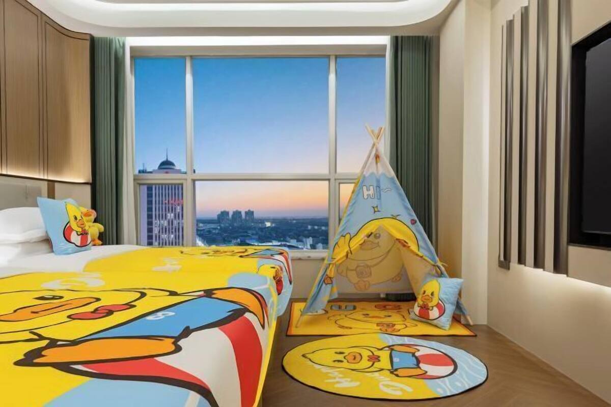 A hotel room with a large bed featuring a cartoon character design, a window with a cityscape view, a teepee with a cartoon character, and a flat-screen TV mounted on the wall.