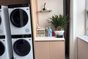 A laundry area with stacked washing machines, a small shelf with decorative items, and a potted plant. - Phoenix Court Hotel (Jingzhou Ancient City Changjiang University) (Jingzhou)
