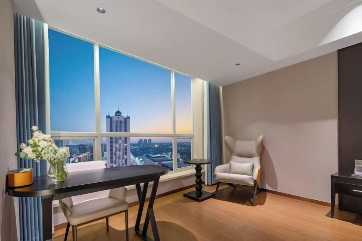A modern hotel room with a large window, a dining table, a chair, and a small side table.