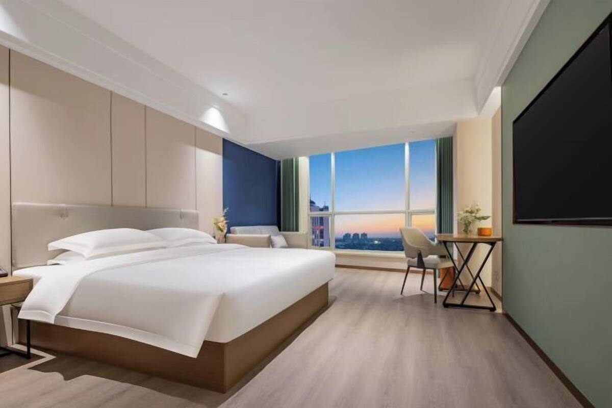 A modern hotel room with a large bed, a desk, and a view of the cityscape.