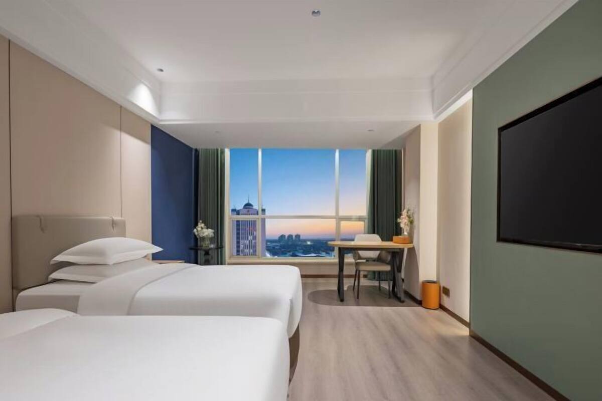 A hotel room with two beds, a desk, a chair, a TV, and a large window with a city view.