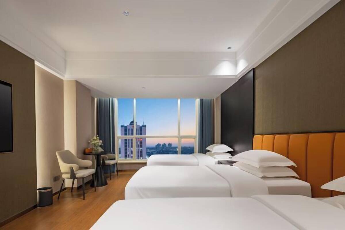 A hotel room with two beds, a desk, a chair, and a large window with a city view.