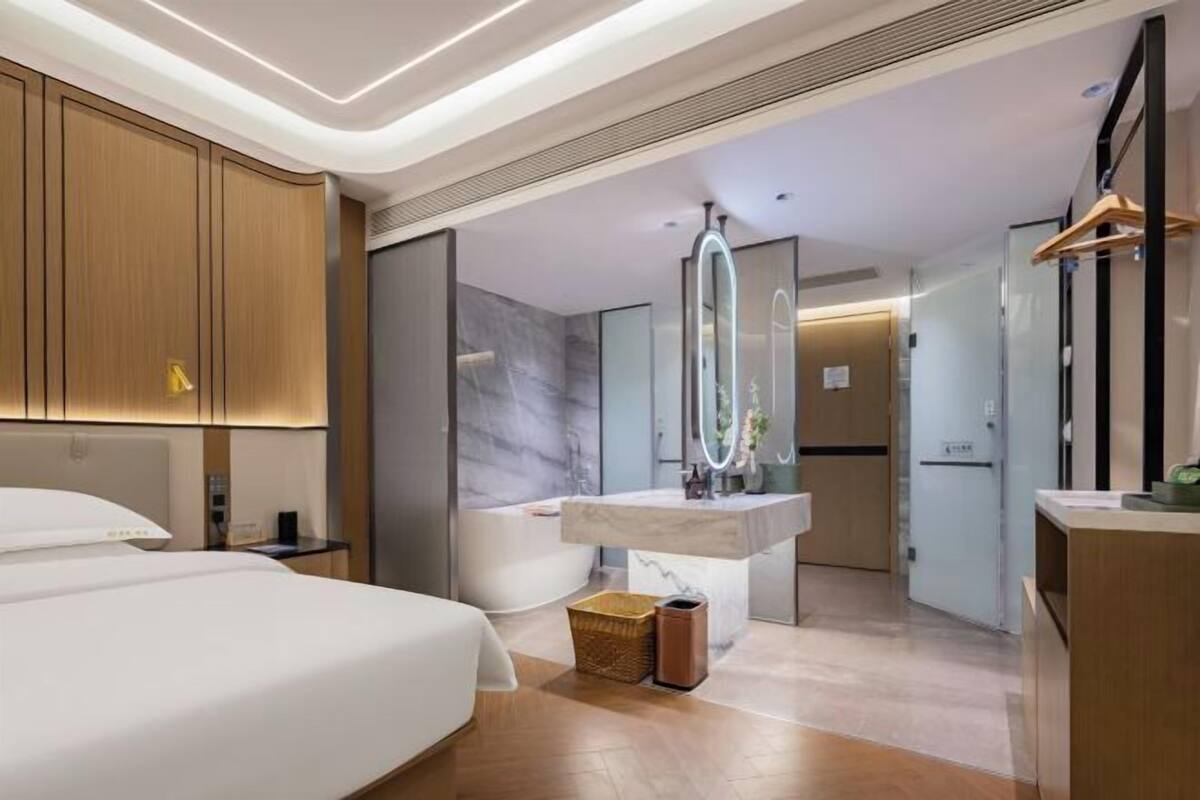 A modern hotel room with a large bed, a bathroom with a bathtub, a vanity with a mirror, and a small seating area.