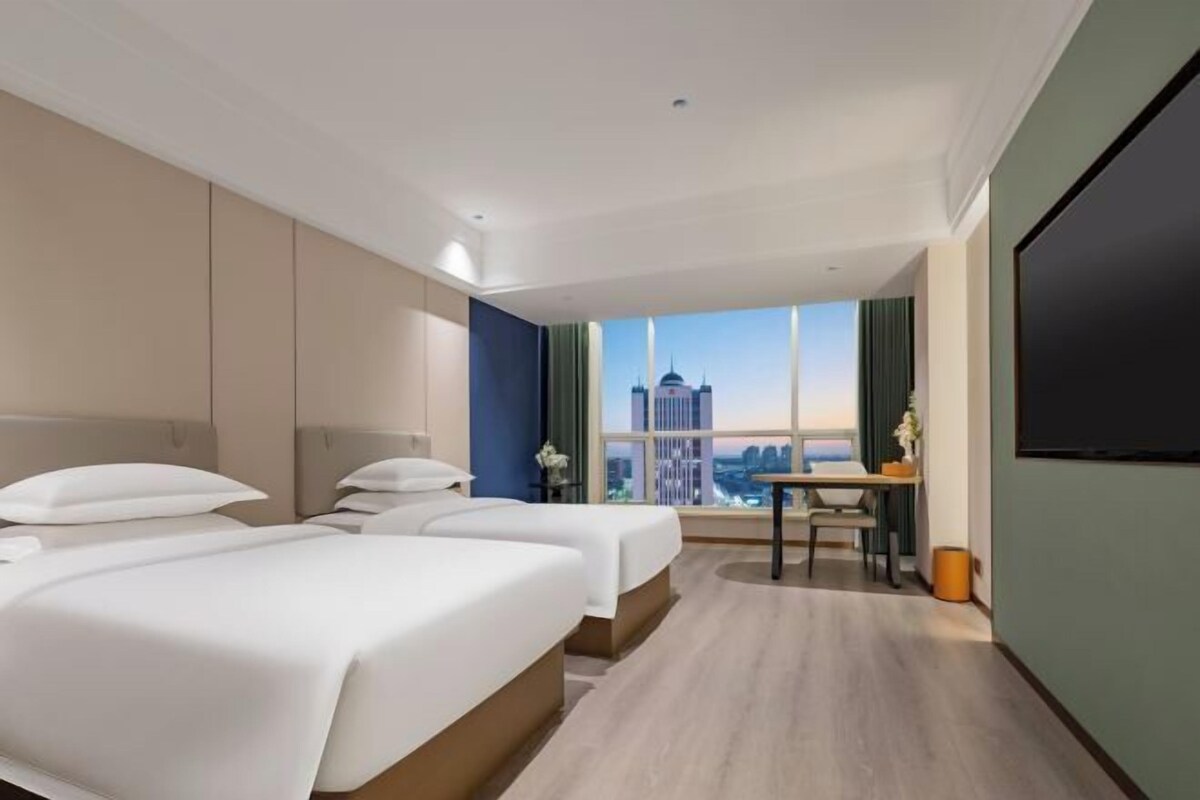 A hotel room with two beds, a desk, a chair, a TV, and a large window with a city view.