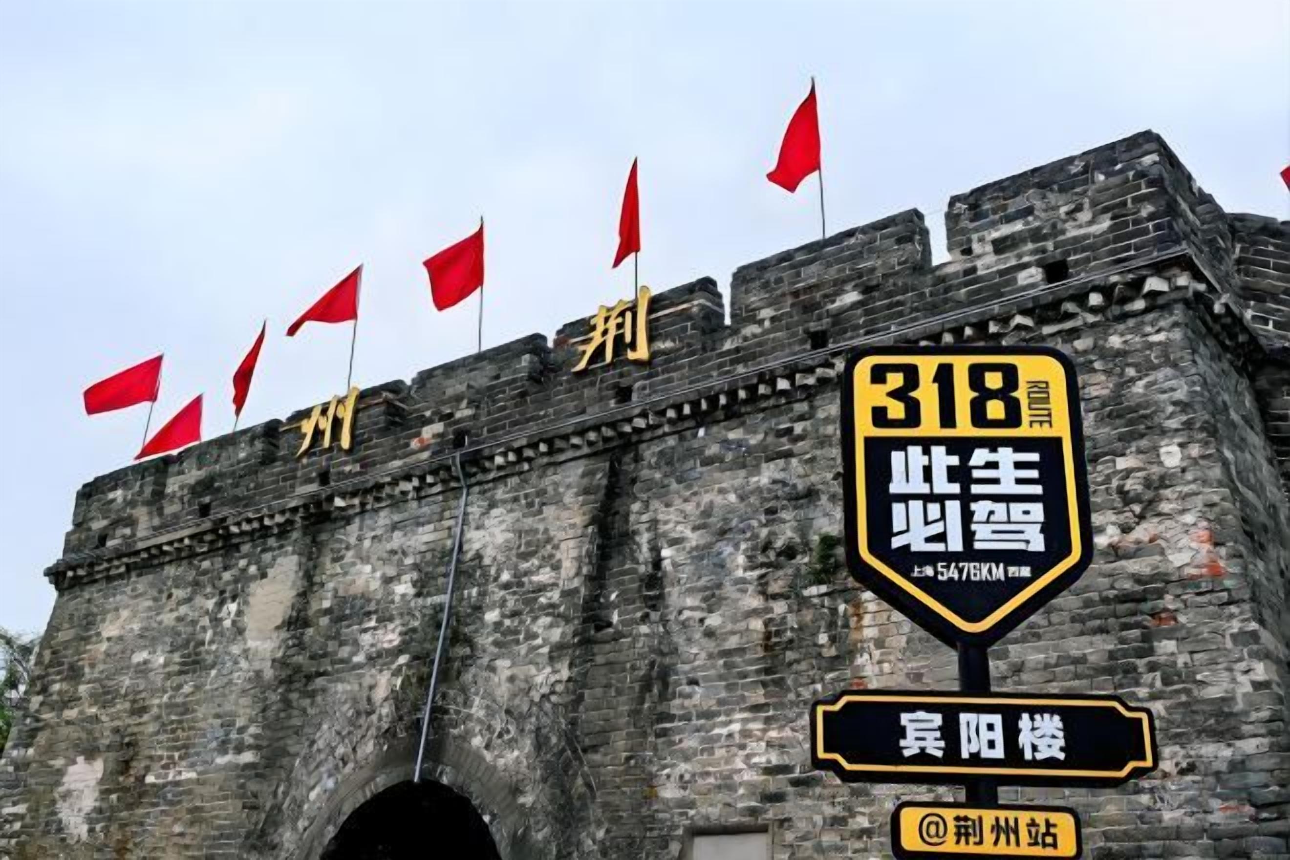 Een historische stenen poort met rode vlaggen, een bord met route 318 en een zwart bord met goudkleurige letters.