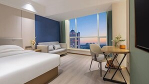 A modern hotel room with a large bed, a sofa, a desk, and a view of a cityscape at sunset. - Phoenix Court Hotel (Jingzhou Ancient City Changjiang University) (Jingzhou)