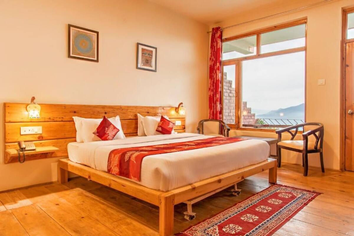 Una cama de madera con ropa de cama blanca y detalles en rojo, una mesita de noche de madera con una lámpara, una silla y una ventana con vistas a montañas.