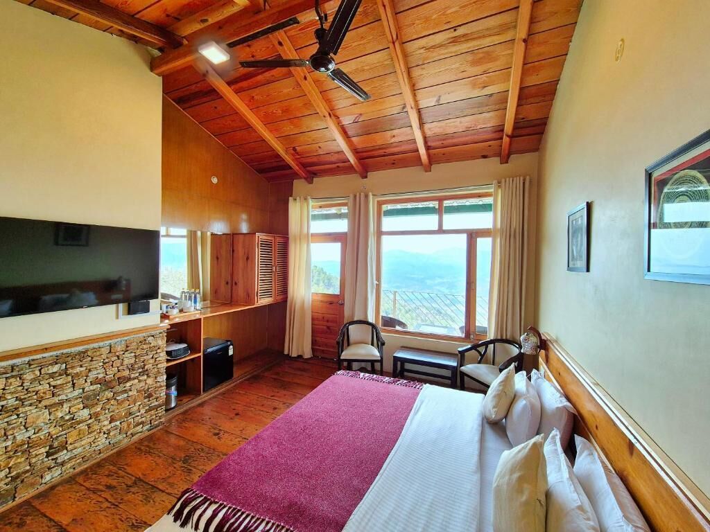 A room with a stone wall, wooden ceiling, a bed with a pink blanket, two chairs, a TV, and a view of the landscape.