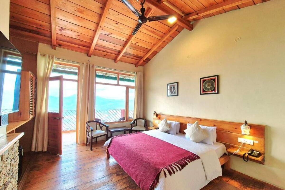 Una habitación con techo de madera, cama con ropa blanca y un plaid rojo, dos sillas, un escritorio con una computadora y una ventana con cortinas.