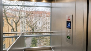 An elevator with a digital display, a handrail, and a window with a view of buildings and trees. - Loft Jecna 43 Prague (Prague)