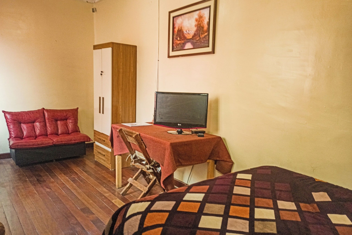 A room with a wooden floor, a red sofa, a desk with a computer, a chair, and a bed with a patterned blanket.
