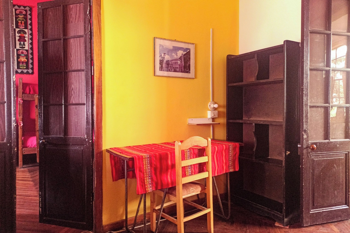 Una habitación con piso de madera, una mesa roja con una silla, una estantería y una puerta.