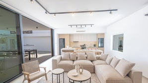 A modern living room with a beige sectional sofa, two round coffee tables, and a wooden chair. A large glass door leads to an outdoor area. - Ayia Napa Orea Villa OR9 (Ayia Napa)
