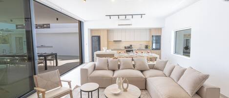 A modern living room with a beige sectional sofa, two round coffee tables, and a wooden chair. A large glass door leads to an outdoor area.