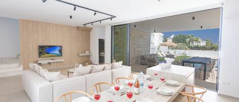 A modern living room with a large TV, a dining area with a table set for four, and a view of the outdoors.