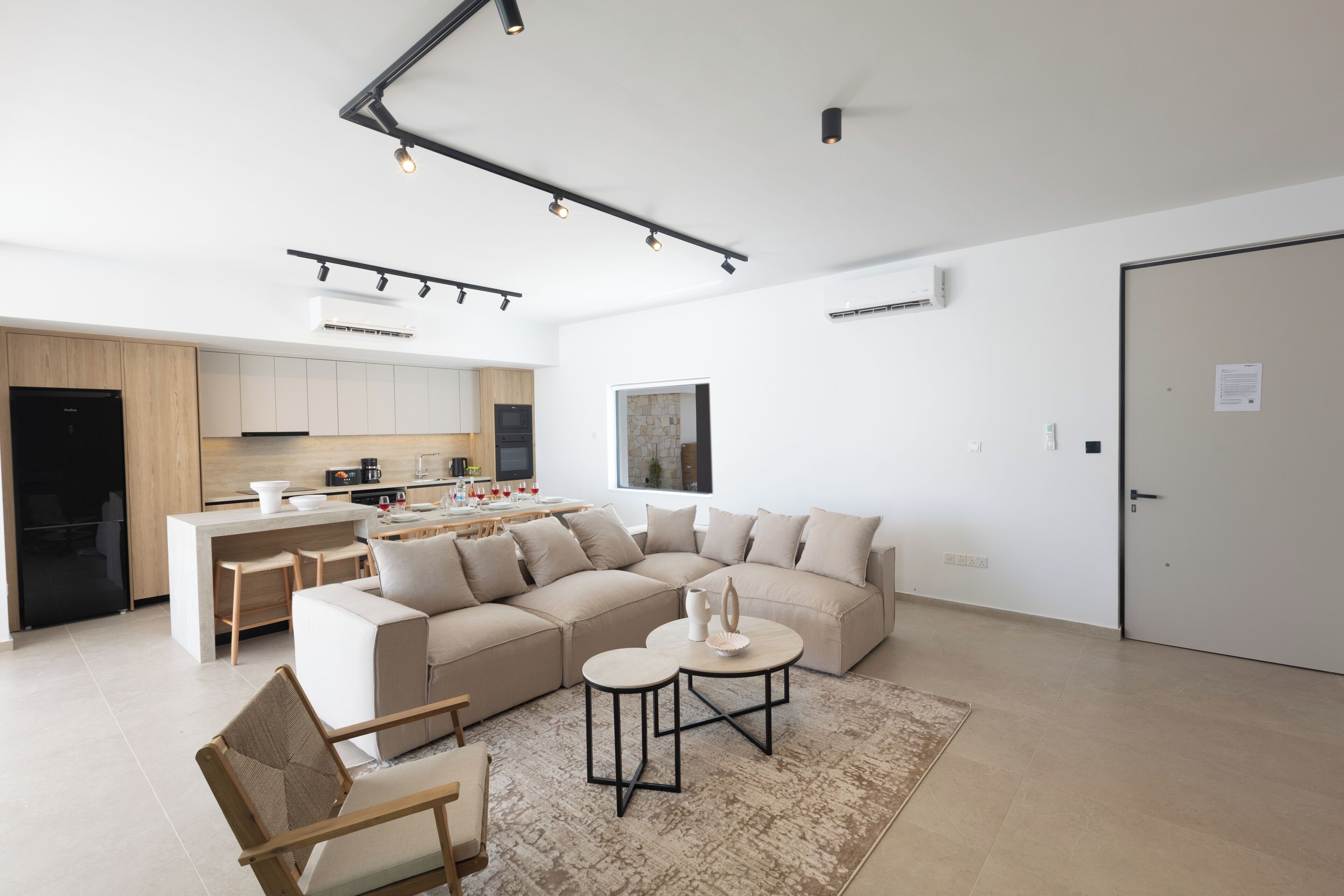 Ein modernes Wohnzimmer mit einem beigefarbenen Ecksofa, einem Couchtisch und einem Essbereich, im Hintergrund befindet sich eine Küche.