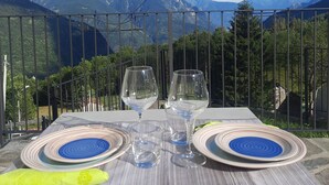 Une table avec deux assiettes et deux verres à vin, dressée sur une nappe, d’où l’on peut admirer un paysage montagneux.