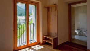 Une pièce dotée d’un plancher en bois, d’un banc en bois, d’une armoire en bois et offrant une vue sur une montagne et des bâtiments, visible à travers une porte vitrée.