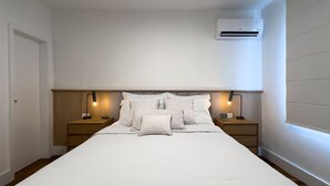 A neatly made bed with white linens and pillows, two bedside tables with lamps, and an air conditioning unit on the wall.