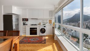 Une cuisine moderne avec des meubles blancs, un four encastré, un lave-linge et une grande fenêtre donnant sur la ville.
