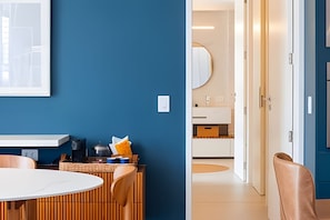 Una habitación moderna con pared azul, una mesa redonda y una silla de madera. Hay una puerta que conduce a un baño con un espejo y un lavabo.